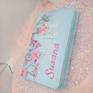 Biblia RVR1960 personalizada en celeste con diseño floral, cierre de zíper, nombre en piedrería e índices de pestañas.